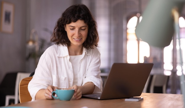 Jonge blanke brunette meisje in shirt zit aan tafel met laptop en kopje koffie binnenshuis