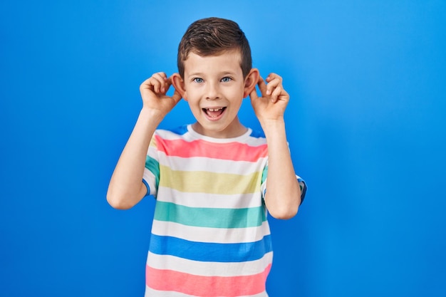 Jonge blanke jongen die over een blauwe achtergrond staat glimlachend oren trekt met vingers, grappig gebaar. auditie probleem
