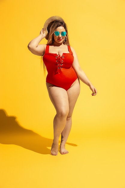 Jonge blanke plus size vrouwelijk model bereidt zich voor op strandresort op gele muur. Vrouw in rode zwembroek, hoed en zonnebril. Concept van zomer, feest, lichaam positief, gelijkheid en chill.