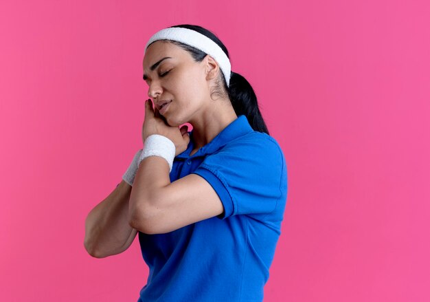 Jonge blanke sportieve vrouw met hoofdband en polsbandjes staat zijwaarts alsof ze op roze met kopie ruimte slaapt