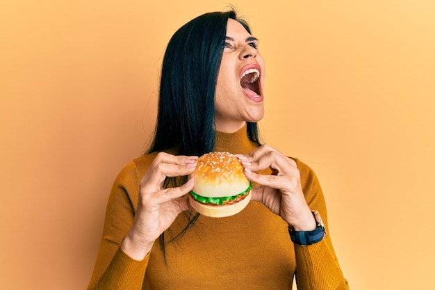 Gratis foto jonge blanke vrouw die een smakelijke klassieke hamburger eet, boos en boos schreeuwend gefrustreerd en woedend schreeuwend met woede die omhoog kijkt