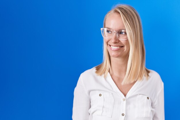 Jonge blanke vrouw die over een blauwe achtergrond staat en wegkijkt met een glimlach op het gezicht, natuurlijke uitdrukking. zelfverzekerd lachen.