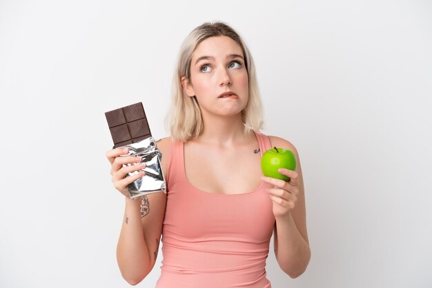 Jonge blanke vrouw geïsoleerd op een witte achtergrond die twijfels heeft terwijl ze een chocoladetablet in de ene hand en een appel in de andere neemt