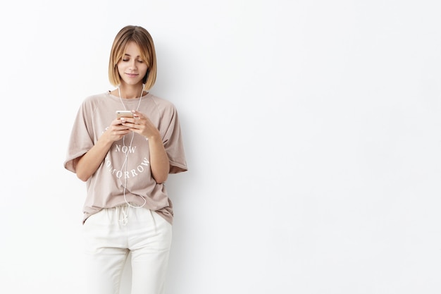 Jonge blanke vrouw met kort kapsel, terloops gekleed, mobiele telefoon in handen houden, berichten typen, naar muziek luisteren met een koptelefoon