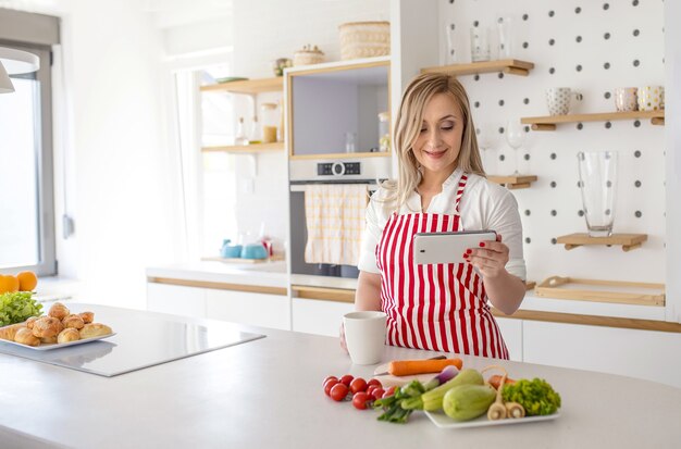 Jonge blanke vrouw met rood gestreepte schort met mok en kijkt naar recept op telefoon in de keuken