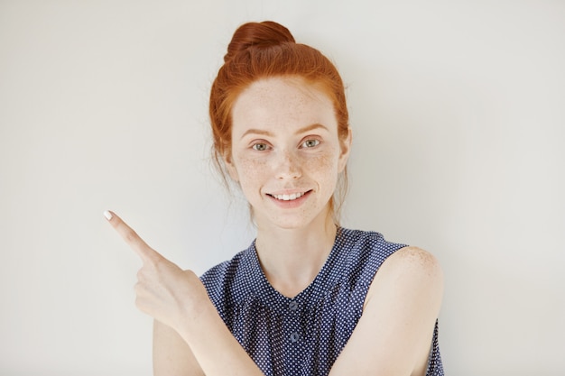 Jonge blanke vrouw met schone sproeten huid en gember haar wijzend haar vinger zijwaarts, iets te demonstreren op witte blinde muur