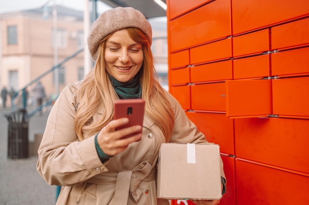 Jonge blanke vrouw met smartphone haalt post op van geautomatiseerde selfservice postterminalmachine