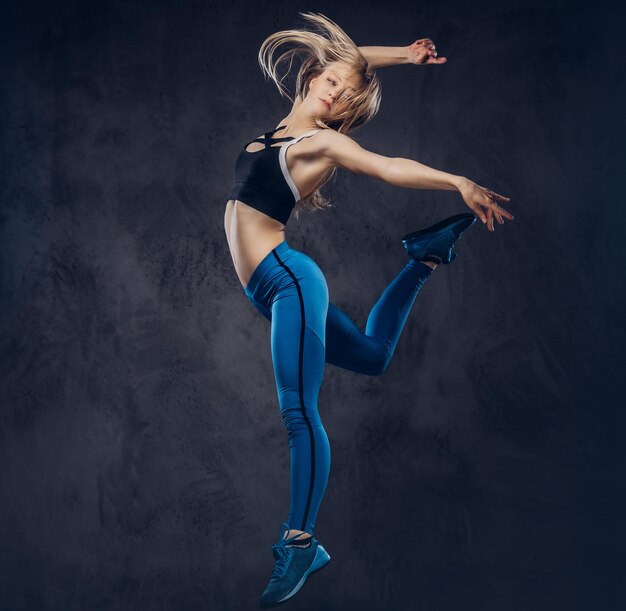 Jonge blonde ballerina in sportkleding danst en springt in een studio. Geïsoleerd op een donkere achtergrond.