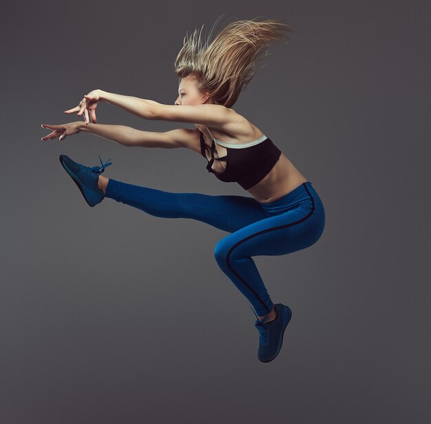 Gratis foto jonge blonde ballerina in sportkleding danst en springt in een studio. geïsoleerd op een grijze achtergrond.