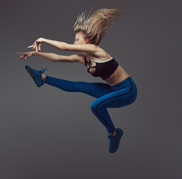 Jonge blonde ballerina in sportkleding danst en springt in een studio. Geïsoleerd op een grijze achtergrond.
