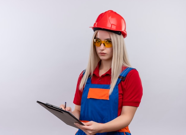 Jonge blonde ingenieur bouwer meisje in uniform dragen veiligheidsbril schrijven op klembord op geïsoleerde witte muur