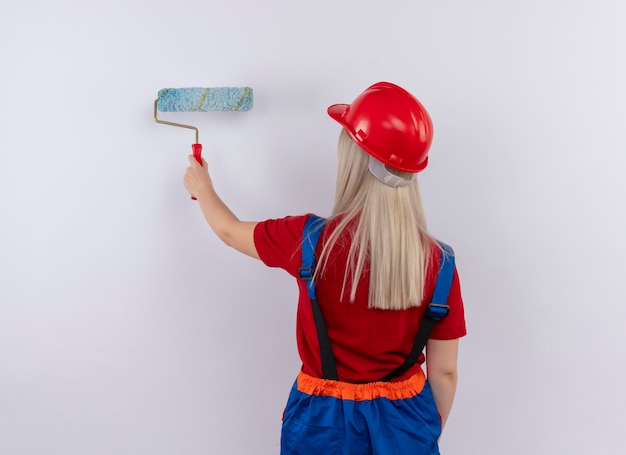 Jonge blonde ingenieur bouwer meisje in uniform schilderij muur met verfroller staande achter zicht op geïsoleerde witte muur