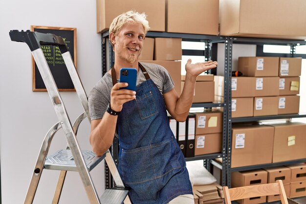 Jonge blonde man met behulp van smartphone werken bij pakhuis glimlachend vrolijk presenteren en wijzen met handpalm kijkend naar de camera