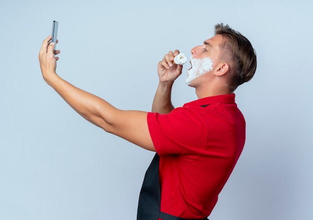 Jonge blonde mannelijke kapper in uniform staat zijwaarts uitstrijkjes scheerschuim op gezicht houden en kijken naar telefoon geïsoleerd op witte ruimte met kopie ruimte