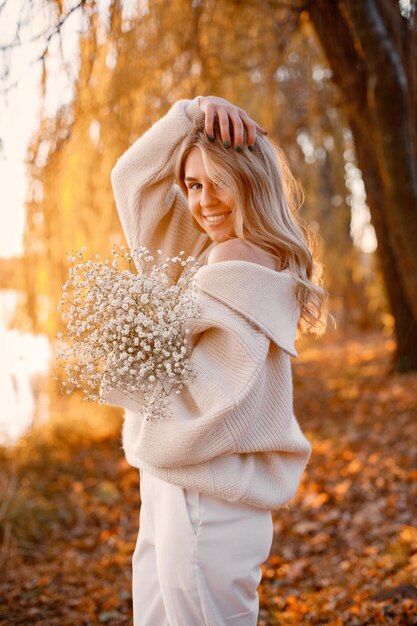 Jonge blonde meid met bloemen die in het herfstpark in de buurt van het meer staat Vrouw draagt beige trui Meisje poseert voor een foto op zonnige dag