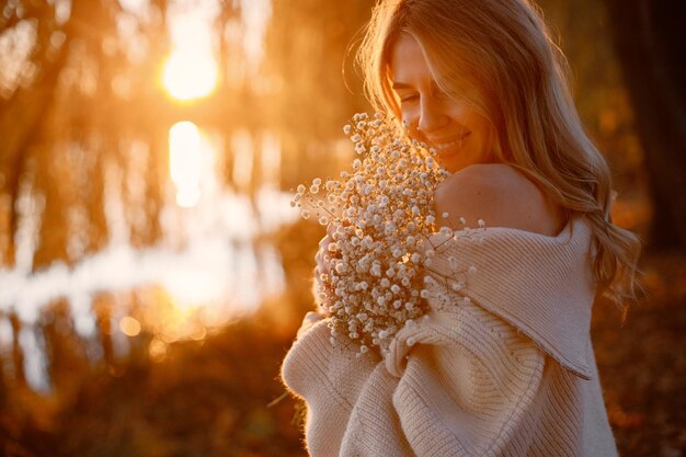 Jonge blonde meid met bloemen die in het herfstpark in de buurt van het meer staat Vrouw draagt beige trui Meisje poseert voor een foto op zonnige dag