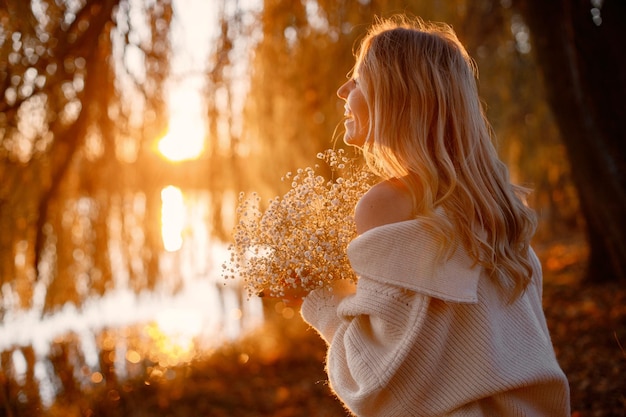 Jonge blonde meid met bloemen die in het herfstpark in de buurt van het meer staat Vrouw draagt beige trui Meisje poseert voor een foto op zonnige dag