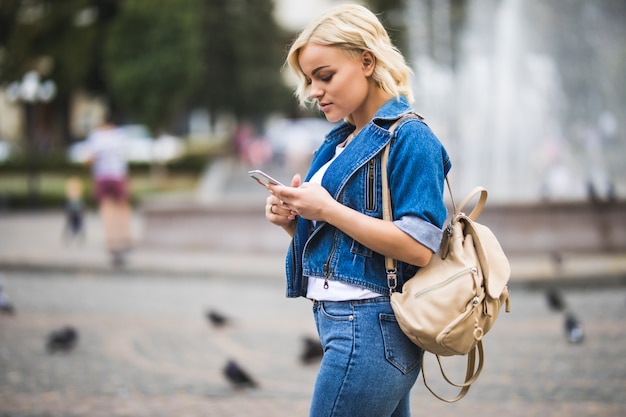 Jonge blonde meisje vrouw met telefoon in haar handen op straat plein fontain gekleed in spijkerbroek suite met tas op haar schouder in zonnige dag