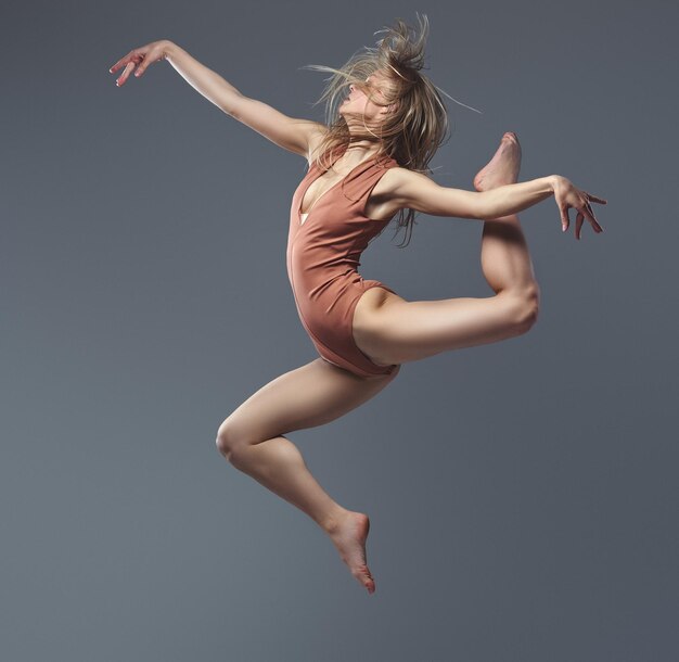 Jonge blonde sierlijke ballerina danst en springt in een studio. Geïsoleerd op een grijze achtergrond.