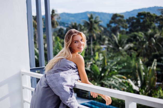 Gratis foto jonge blonde stijlvolle europese vrouw in deken op tropisch balkon ontmoet zonsopgang in de ochtend.