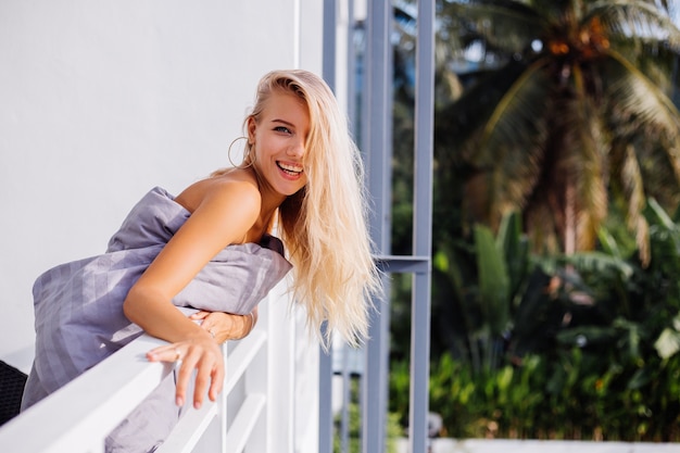 Jonge blonde stijlvolle europese vrouw in deken op tropisch balkon ontmoet zonsopgang in de ochtend.