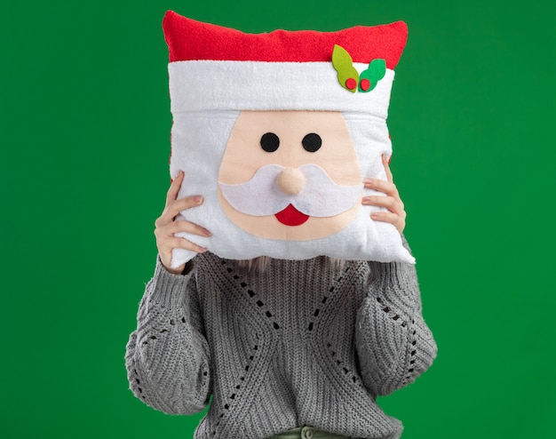 Jonge blonde vrouw die in de wintertrui Kerstmishoofdkussen houdt dat haar gezicht met hoofdkussen verbergt dat zich over groene achtergrond bevindt