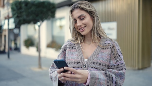 Jonge blonde vrouw die lacht zelfverzekerd met behulp van smartphone op straat