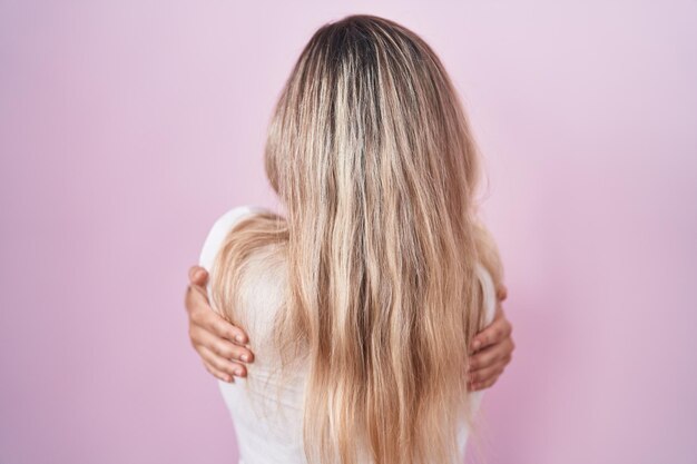 Jonge blonde vrouw die over een roze achtergrond staat en zichzelf gelukkig en positief knuffelt van achterwaartse zelfliefde en zelfzorg