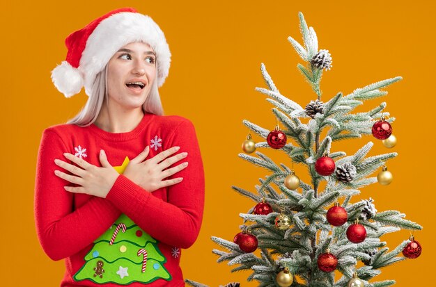 Jonge blonde vrouw in kerst trui en kerstmuts opzij kijken hand in hand op haar borst hapy en positief staan naast een kerstboom op oranje achtergrond