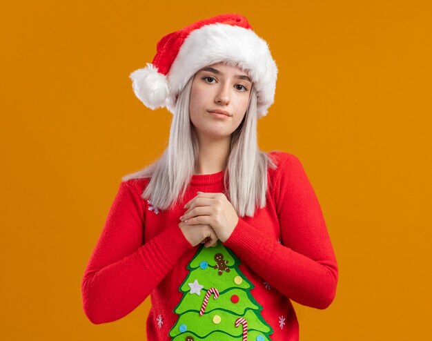 Jonge blonde vrouw in Kerstmissweater en kerstmuts kijken camera met ernstige gezicht hand in hand samen permanent over oranje achtergrond