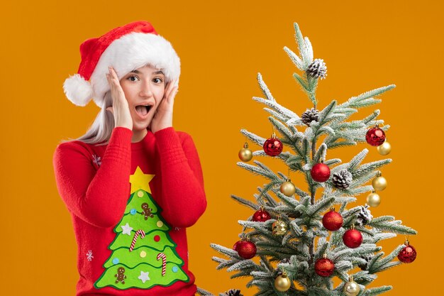 Jonge blonde vrouw in Kerstmissweater en santahoed verbaasd en verrast
