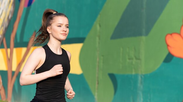 Jonge blonde vrouw in sportkleding draait op de weg bij buiten training, veelkleurige muur op de achtergrond