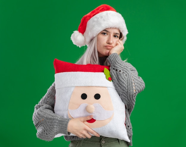 jonge blonde vrouw in winter trui en kerstmuts bedrijf kerst kussen kijken camera wordt verward en ontevreden staande over groene achtergrond