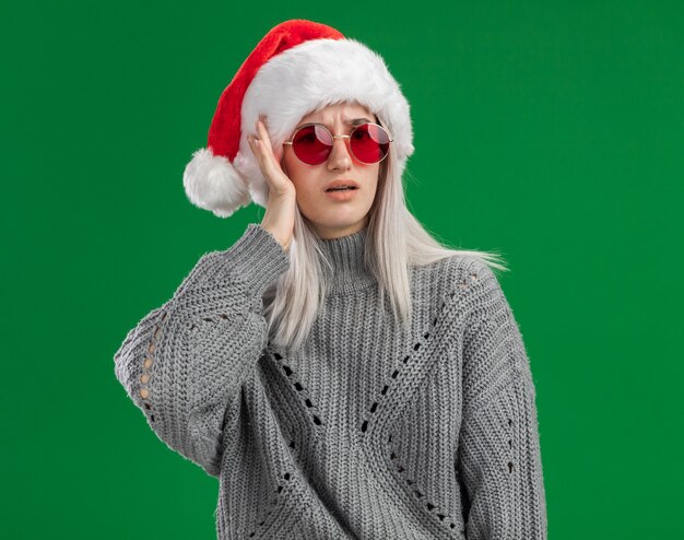 Jonge blonde vrouw in winter trui en kerstmuts met rode bril op zoek verward met de hand op haar hoofd staande over groene achtergrond