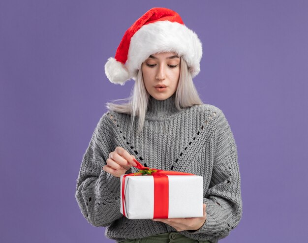 Jonge blonde vrouw in wintertrui en kerstmuts die een cadeautje vasthoudt om het te openen omdat ze geïntrigeerd is en over een paarse muur staat