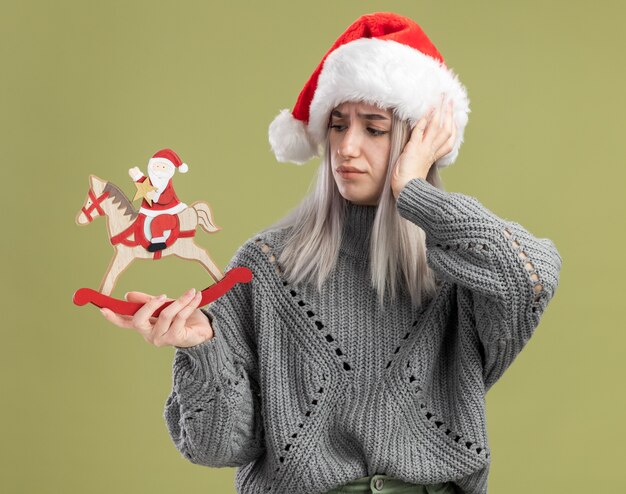 Jonge blonde vrouw in wintertrui en kerstmuts die kerstspeelgoed vasthoudt en ernaar kijkt verward met de hand op haar hoofd die over de groene muur staat