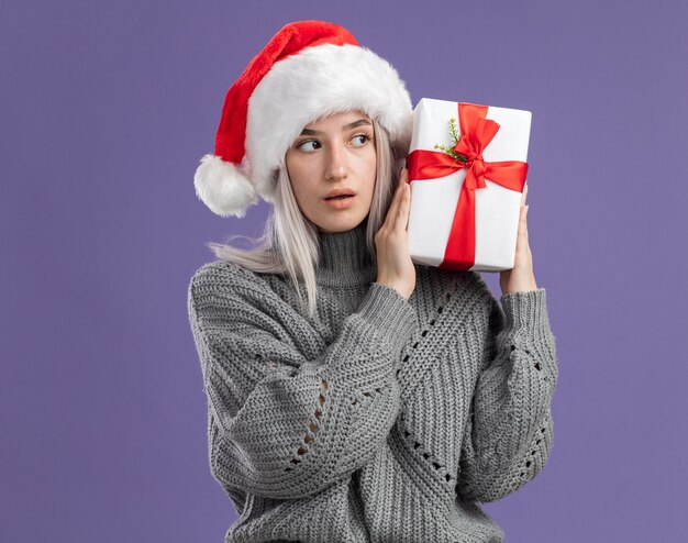 Jonge blonde vrouw in wintertrui en kerstmuts met een cadeautje en kijkt geïntrigeerd over een paarse muur