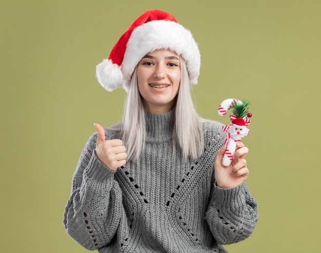 Jonge blonde vrouw in wintertrui en kerstmuts met kerstsnoepgoed glimlachend vrolijk duimen omhoog staande over groene muur