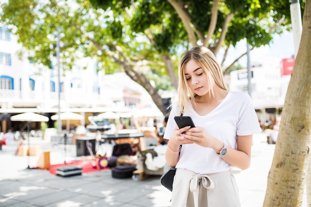 Jonge blonde vrouw lopen en typen op telefoon in de straat op een zonnige zomerdag