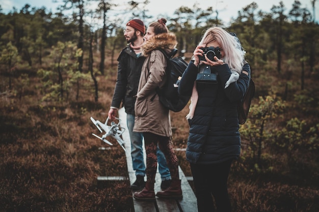 Jonge blonde vrouw maakt een foto met haar camera terwijl ze met haar vrienden in het bos wandelt.