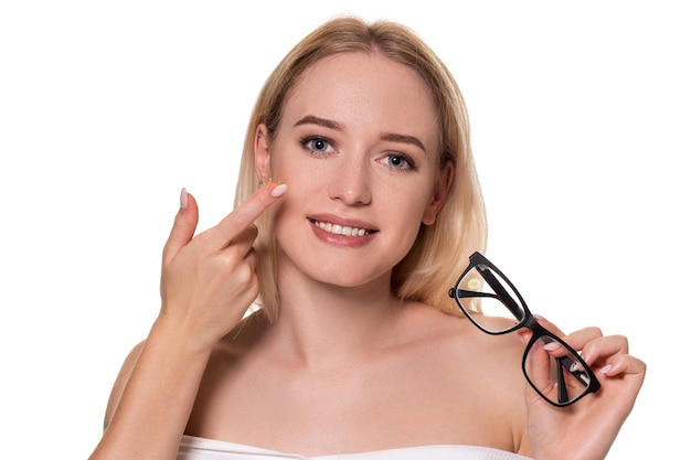 Jonge blonde vrouw met contactlens op vinger voor haar gezicht en in haar andere hand een zwarte bril op witte achtergrond. Gezichtsvermogen en oogzorgconcept. Concept van keuze