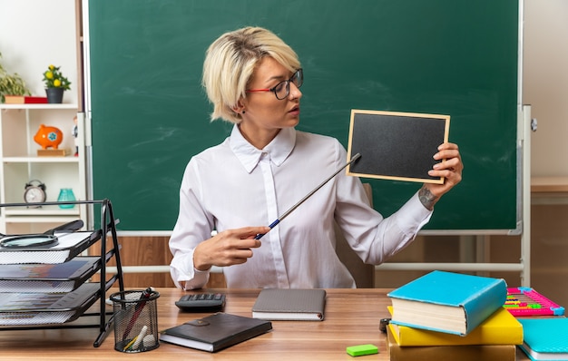 Jonge blonde vrouwelijke leraar met een bril die aan een bureau zit met schoolhulpmiddelen in de klas met een mini-bord dat ernaar kijkt en ernaar wijst met een aanwijzer