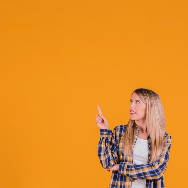 Jonge blondevrouw die haar vinger richten die omhoog tegen een oranje achtergrond omhoog kijken