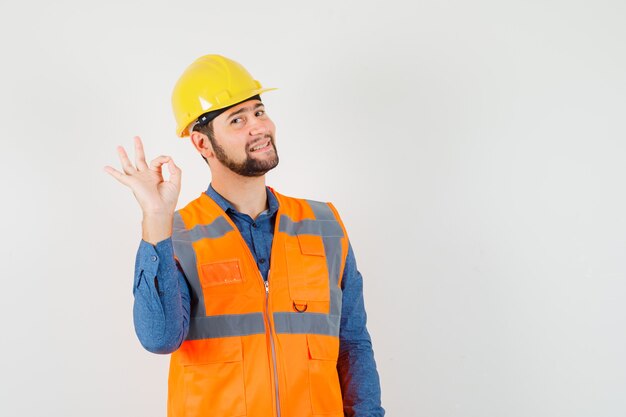 Jonge bouwer die ok gebaar in overhemd, vest, helm toont en vrolijk, vooraanzicht kijkt.