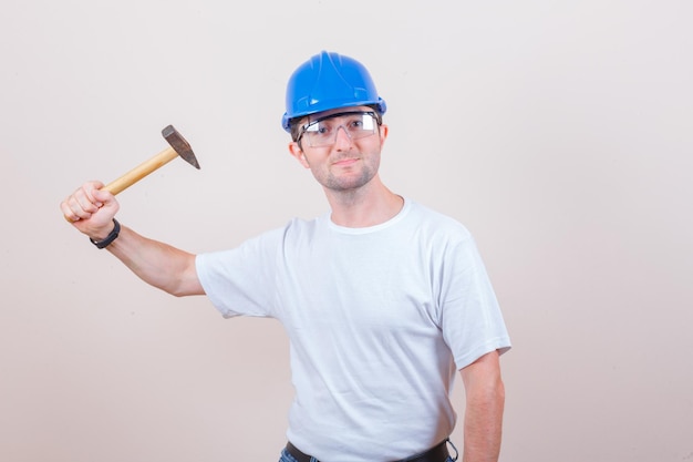 Jonge bouwer dreigt met hamer in t-shirt, helm en kijkt geamuseerd