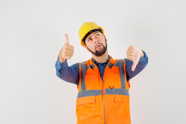 Jonge bouwer in overhemd, vest, helm die duimen op en neer toont en aarzelend, vooraanzicht kijkt.