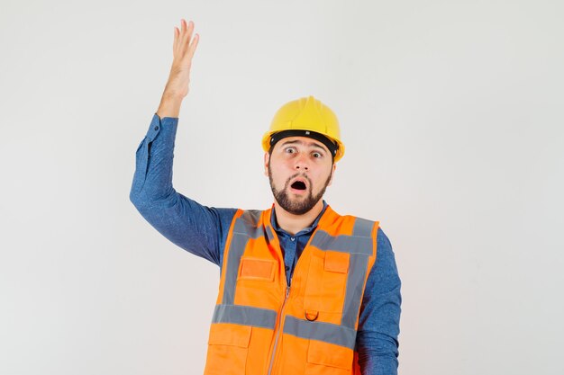 Jonge bouwer in overhemd, vest, helm die hand opheft en geschokt, vooraanzicht kijkt.