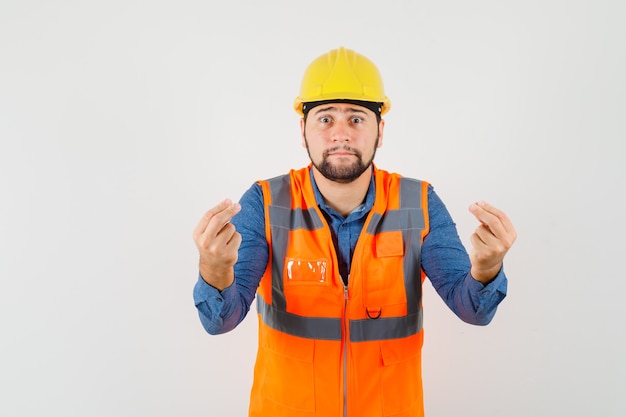 Jonge bouwer in overhemd, vest, helm die iets proberen uit te leggen en verontrust, vooraanzicht kijkt.