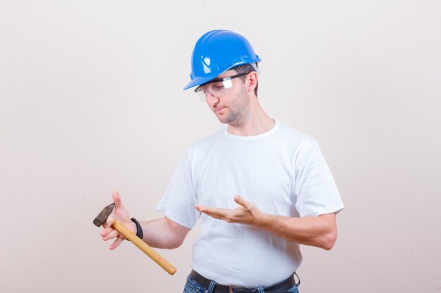 Jonge bouwer in t-shirt, jeans, helm die hamer toont en peinzend kijkt looking