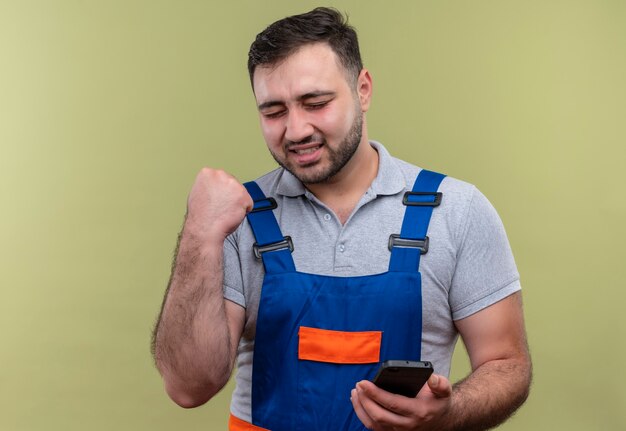 Jonge bouwer man in bouw uniform bedrijf smartphone balde vuist blij en verlaten